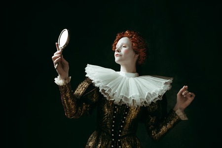 Proud of her self. Medieval redhead young woman in golden vintage clothing as a duchess looking in the mirror on dark green background. Concept of comparison of eras, modernity and renaissance.の写真素材