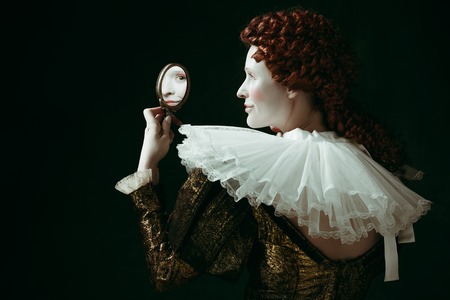 Whos that pretty. Medieval redhead young woman in golden vintage clothing as a duchess looking in the mirror on dark green background. Concept of comparison of eras, modernity and renaissance.の写真素材