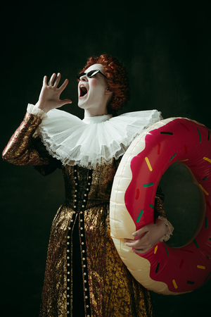 Adventures cant wait. Medieval redhead young woman in golden vintage clothing as a duchess standing on dark green background. Concept of comparison of eras, modernity and renaissance, summer, chill.の写真素材