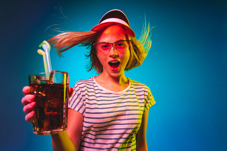 Beautiful female half-length portrait isolated on blue neon lights studio background. Young woman in red cap and sunglasses with drink. Facial expression, summer, weekend concept. Trendy colors.の写真素材