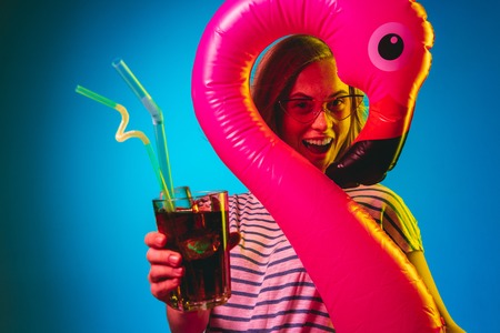 Beautiful female half-length portrait on blue neon lights studio background. Woman in red sunglasses and rubber ring as a flamingo with drink. Facial expression, summer, weekend concept. Trendy colors.の写真素材