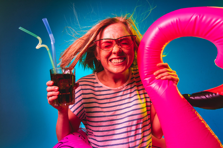 Beautiful female half-length portrait on blue neon lights studio background. Woman in red sunglasses and rubber ring as a flamingo with drink. Facial expression, summer, weekend concept. Trendy colors.の写真素材