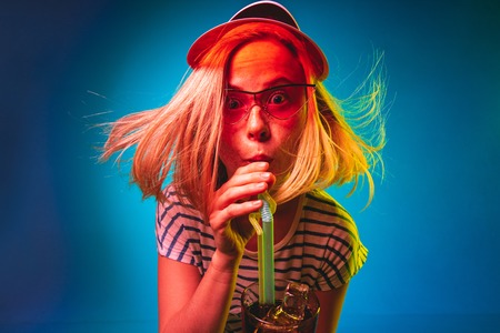 Beautiful female half-length portrait isolated on blue neon lights studio background. Young woman in red cap and sunglasses with drink. Facial expression, summer, weekend concept. Trendy colors.の写真素材