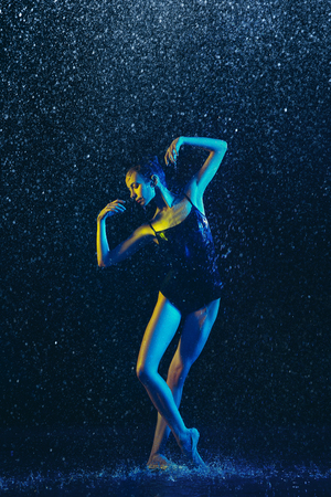 Young female ballet dancer performing under water drops and spray. Caucasian model dancing in neon lights. Attractive woman. Ballet and contemporary choreography concept. Creative art photo.の写真素材