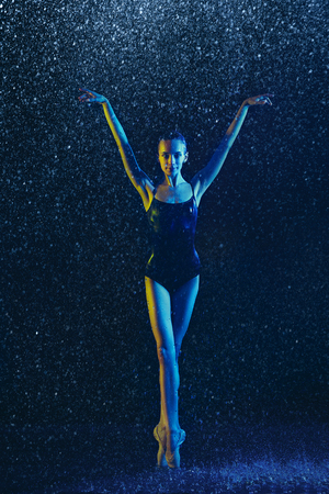 Young female ballet dancer performing under water drops and spray. Caucasian model dancing in neon lights. Attractive woman. Ballet and contemporary choreography concept. Creative art photo.の写真素材