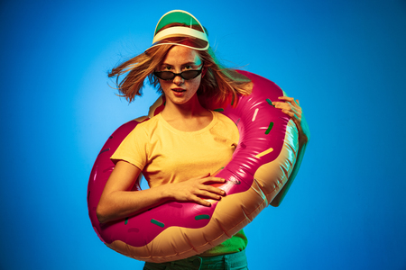 Beautiful female half-length portrait isolated on blue neon lights studio background. Woman in green cap, rubber ring and sunglasses. Facial expression, summer, weekend concept. Trendy colors.の写真素材