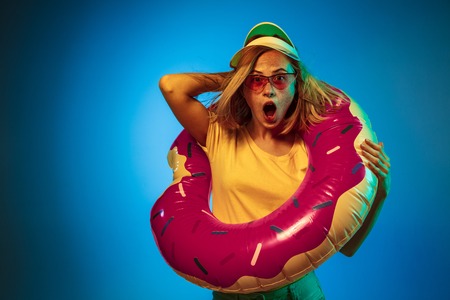 Beautiful female half-length portrait isolated on blue neon lights studio background. Woman in green cap, rubber ring and sunglasses. Facial expression, summer, weekend concept. Trendy colors.の写真素材