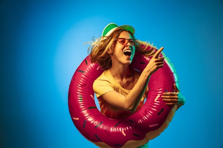 Beautiful female half-length portrait isolated on blue neon lights studio background. Woman in green cap, rubber ring and sunglasses. Facial expression, summer, weekend concept. Trendy colors.の写真素材