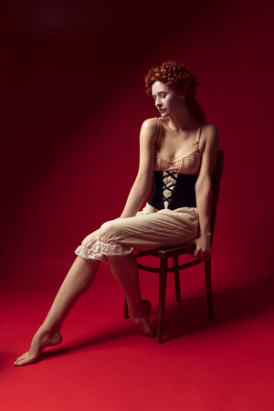 Medieval redhead young woman as a duchess in black corset and night clothes sitting on the chair on redの写真素材