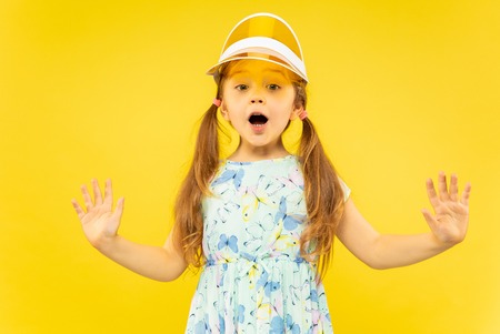 Beautiful emotional little girl isolated on yellow background. Half-lenght portrait of happy and astonished child wearing a dress and orange cap. Concept of summer, human emotions, childhood.の写真素材
