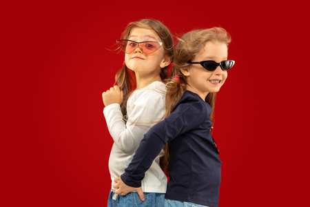 Beautiful emotional little girl isolated on red background. Half-lenght portrait of happy sisters or friends in red and black sunglasses. Concept of facial expression, human emotions, childhood.の写真素材