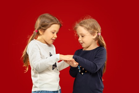 Beautiful emotional little girl isolated on red studio background. Half-lenght portrait of happy sisters or friends standing and smiling. Concept of facial expression, human emotions, childhood.の写真素材