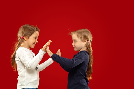 Beautiful emotional little girl isolated on red studio background. Half-lenght portrait of happy sisters or friends standing and smiling. Concept of facial expression, human emotions, childhood.の写真素材