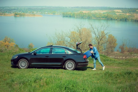 Handsome smiling man in jeans, jacket and sunglasses going to vacations, loading his suitcase in car trunk on the rivers side. Preparing for weekend trip. Concept of summertime, resort, chilling.の写真素材