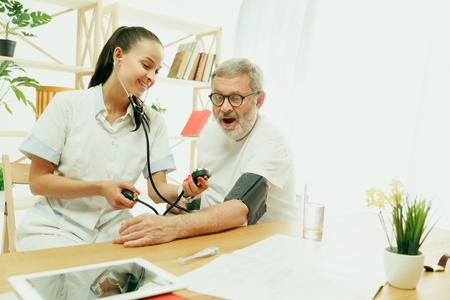 The visiting nurse or health visitor taking care of senior man. Lifestyle portrait at home. Medicine, healthcare and prevention. Girl checking or measuring patients blood pressure during the visit.の写真素材