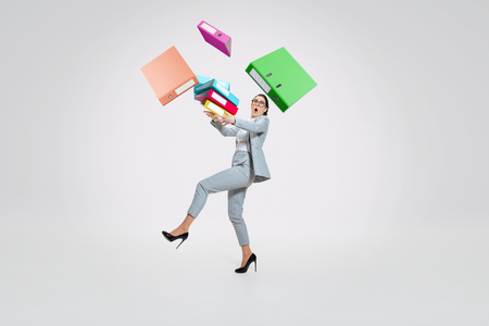 Young woman in suit getting a lot of work and losing concentration. Dropping folders with papers on the go, trying to catch that. Concept of office workers troubles, business, problems and stress.の写真素材