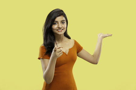 Beautiful female half-length portrait isolated on yellow studio background. Young emotional indian woman in dress pointing and showing. Negative space. Facial expression, human emotions concept.の写真素材