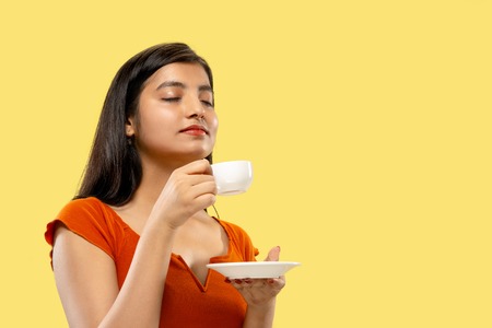 Beautiful female half-length portrait isolated on yellow studio background. Young emotional indian woman in dress drinking coffee. Negative space. Facial expression, human emotions concept.の写真素材