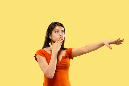 Beautiful female half-length portrait isolated on yellow studio background. Young emotional indian woman in dress pointing and showing. Negative space. Facial expression, human emotions concept.の写真素材