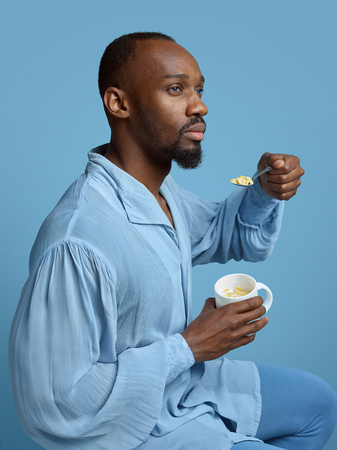 Young man as a medieval grandee on blue background. Portrait of male african-american model in retro shirt. Eating icecream in a cap. Human emotions, comparison of eras, facial expressions concept.の写真素材
