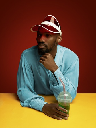 Young man as a medieval grandee or nobleman on red studio background. Holding a glass of drink. Portrait in retro costume and a cap. Human emotions, comparison of eras, facial expressions concept.の写真素材
