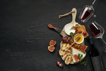 Two glasses of red wine and a tasty cheese plate with fruit, grape, nuts and toasted bread on a...