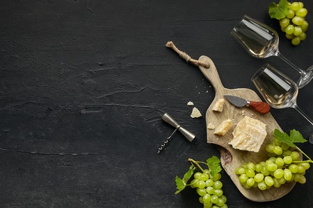 Two glasses of white wine and a tasty cheese plate with fruit on a wooden kitchen plate on the black stone background, top view, copy space. Gourmet food and drink.の写真素材