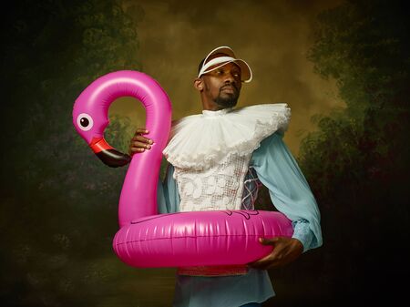 Young man as a medieval grandee or nobleman in a swimring as a pink flamingo on dark studio background. Portrait in retro costume. Human emotions, comparison of eras and facial expressions concept.の写真素材