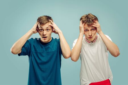 Two young men standing in sportwear isolated on blue studio background. Fans of sport, football or...