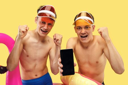 Two young mens half-length portrait isolated on yellow studio background. Smiling friends in caps with swimrings using smartphone. Facial expression, summer, weekend, betting or online paying concept.の写真素材