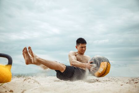 Young healthy man athlete doing exercise with the weight and ball at the beach. Signle male model shirtless training at the river side. Concept of healthy lifestyle, sport, fitness, bodybuilding.の写真素材