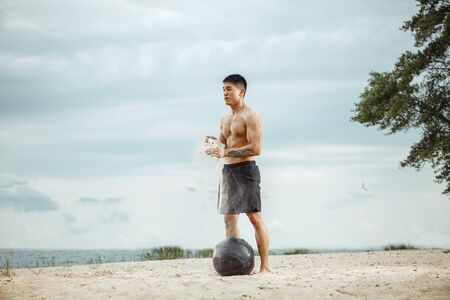 Young healthy man athlete doing exercise with ball at the beach. Signle male model shirtless training air at the river side in sunny day. Concept of healthy lifestyle, sport, fitness, bodybuilding.の写真素材