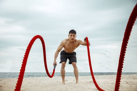 Young healthy man athlete doing exercise with the rope at the beach. Signle male model shirtless training air at the river side in sunny day. Concept of healthy lifestyle, sport, fitness, bodybuilding.の写真素材