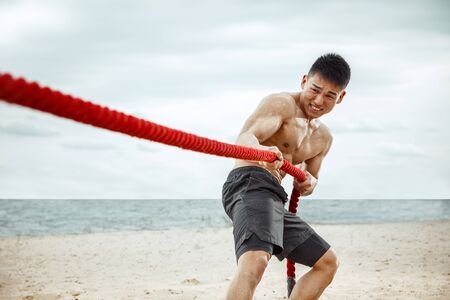 Young healthy man athlete doing exercise with the rope at the beach. Signle male model shirtless training air at the river side in sunny day. Concept of healthy lifestyle, sport, fitness, bodybuilding.の写真素材