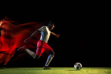Young african-american male football or soccer player in sportwear and boots kicking ball for the goal in mixed light on dark background. Concept of healthy lifestyle, professional sport, hobby.の写真素材
