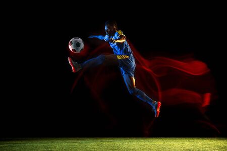 Young african-american male football or soccer player in sportwear and boots kicking ball for the goal in mixed light on dark background. Concept of healthy lifestyle, professional sport, hobby.の写真素材