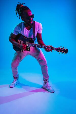 Young african-american musician playing the guitar like a rockstar on blue studio background in neon light. Concept of music, hobby. Joyful attractive guy improvising. Retro colorful portrait.の写真素材