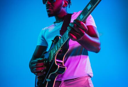 Young african-american musician playing the guitar like a rockstar on blue studio background in neon...
