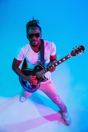 Young african-american musician playing the guitar like a rockstar on blue studio background in neon light. Concept of music, hobby. Joyful attractive guy improvising. Retro colorful portrait.の写真素材