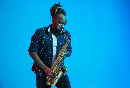 Young african-american jazz musician playing the saxophone on blue studio background in trendy neon light. Concept of music, hobby. Joyful attractive guy improvising. Retro colorful portrait of artist.の写真素材