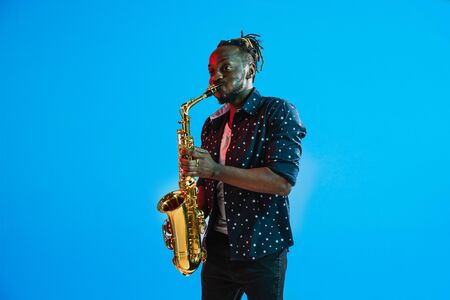 Young african-american jazz musician playing the saxophone on blue studio background in trendy neon light. Concept of music, hobby. Joyful attractive guy improvising. Retro colorful portrait of artist.の写真素材