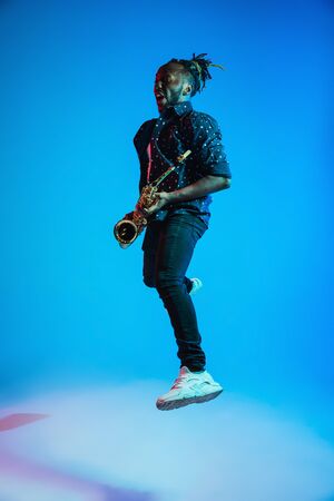 Young african-american jazz musician playing the saxophone on blue studio background in trendy neon light. Concept of music, hobby. Joyful attractive guy improvising. Retro colorful portrait of artist.の写真素材