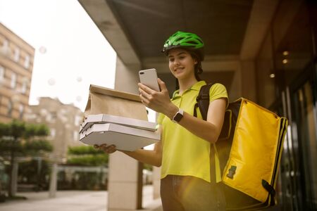 Young woman in yellow shirt delivering pizza using gadgets to track order at the citys street. Courier using online app for receiving payment and tracking shipping address. Modern technologies.の写真素材