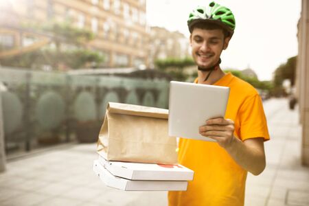 Young man in yellow shirt delivering pizza using gadgets to track order at the citys street. Courier...