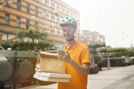 Young man in yellow shirt delivering pizza using gadgets to track order at the citys street. Courier using online app for receiving payment and tracking shipping address. Modern technologies.の写真素材