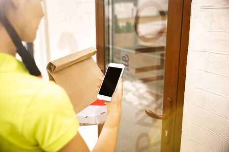 Young woman in yellow shirt delivering pizza using gadgets to track order at the citys street. Courier using online app for receiving payment and tracking shipping address. Modern technologies.の写真素材