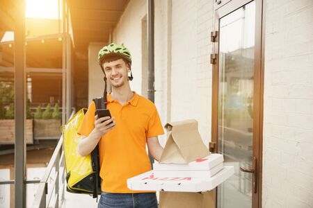 Young man in yellow shirt delivering pizza using gadgets to track order at the citys street. Courier using online app for receiving payment and tracking shipping address. Modern technologies.の写真素材