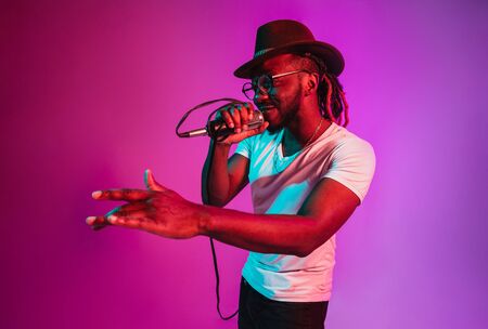 Young african-american jazz musician with microphone singing a song on purple studio background in trendy neon light. Concept of music, hobby, inspirness. Colorful portrait of joyful attractive artist.の写真素材