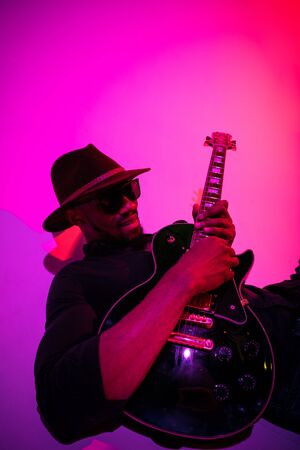 Young african-american musician playing the guitar like a rockstar on gradient purple-pink...