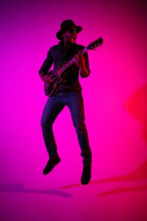 Young african-american musician playing the guitar like a rockstar on gradient purple-pink background in neon light. Concept of music, hobby. Joyful attractive guy improvising. Colorful portrait.の写真素材
